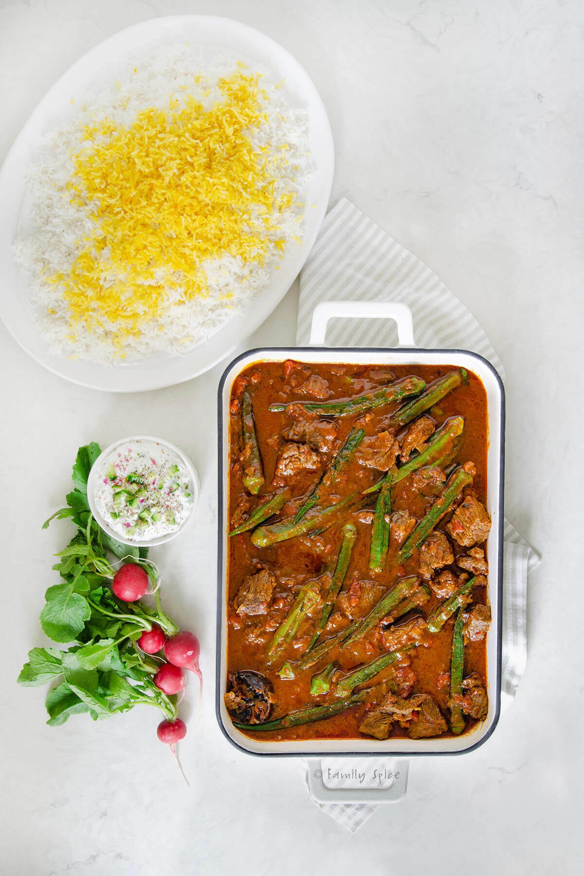 A casserole dish with Persian okra stew in it along with a platter of Persian rice, fresh radishes and a small bowl of yogurt.