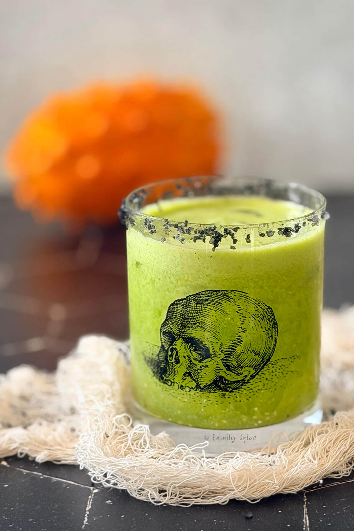 Closeup of a glass with a skull on it with green kiwano halloween margarita in it and charcoal salt on the rim.