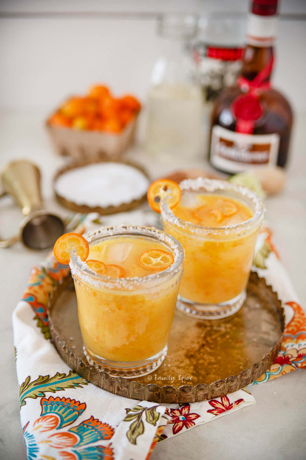 Closeup of two glasses filled with kumquat margaritas on a gold tray with ingredients behind it.