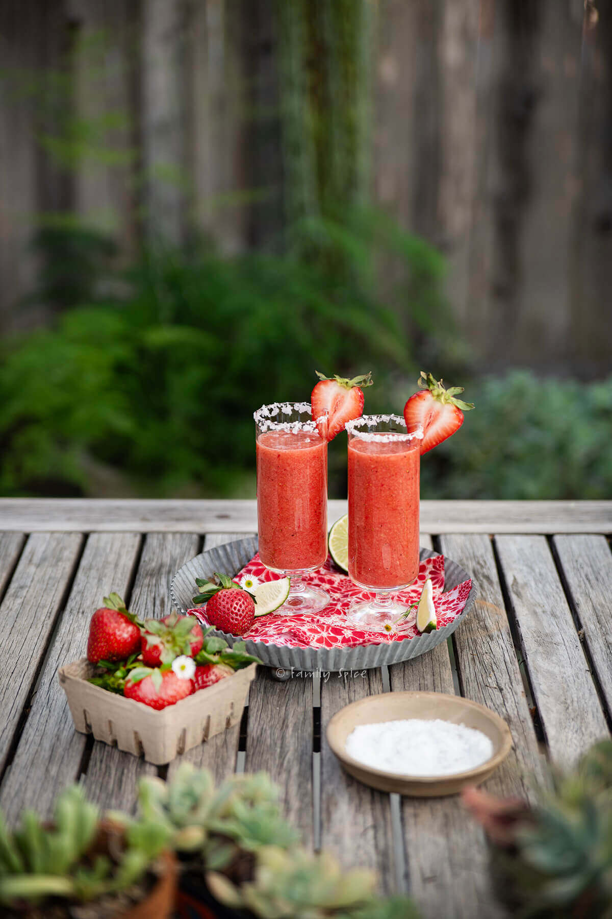 A metal tray with two glasses with strawberry salty dog on an outdoor wood table in a yard.