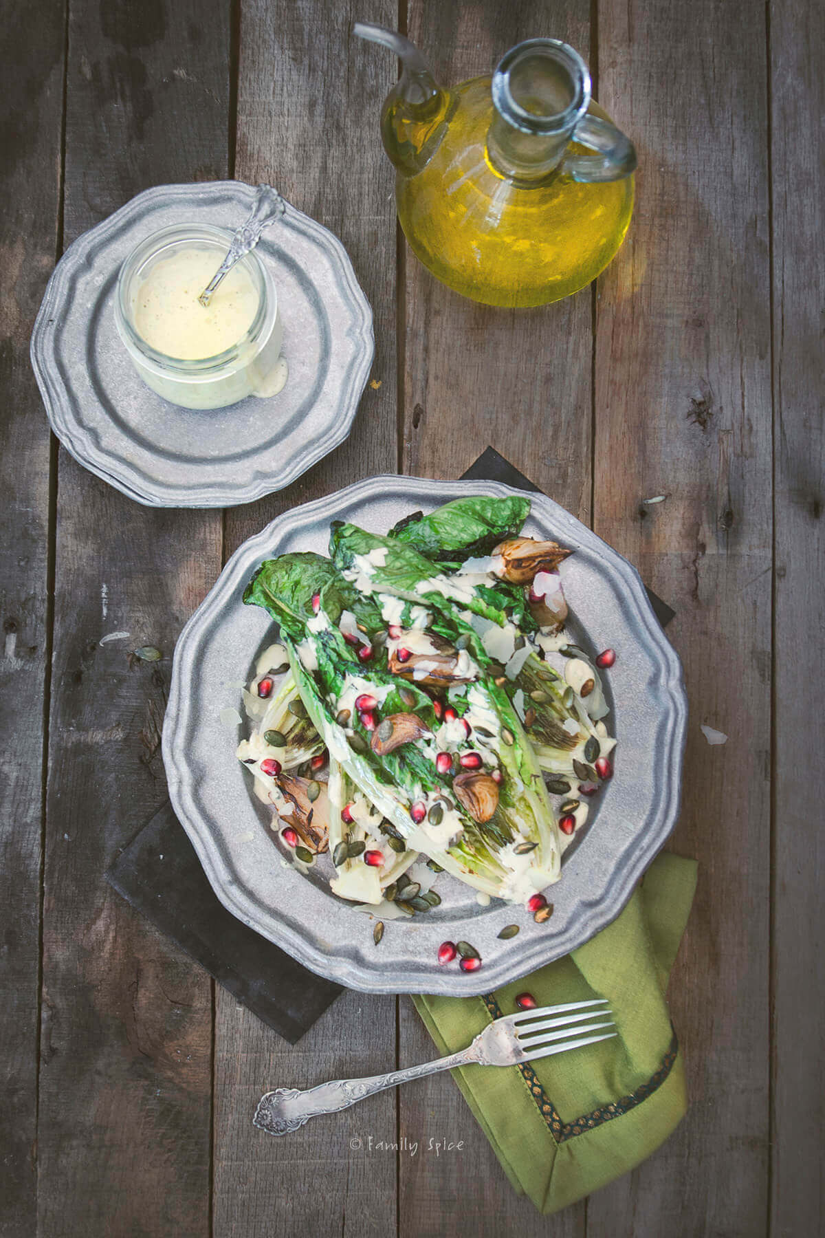 Overhead view of a pewter plate with grilled romaine lettuce topped with roasted shallots and pomegranate arils drizzled with Caesar dressing with a bottle of dressing next to it