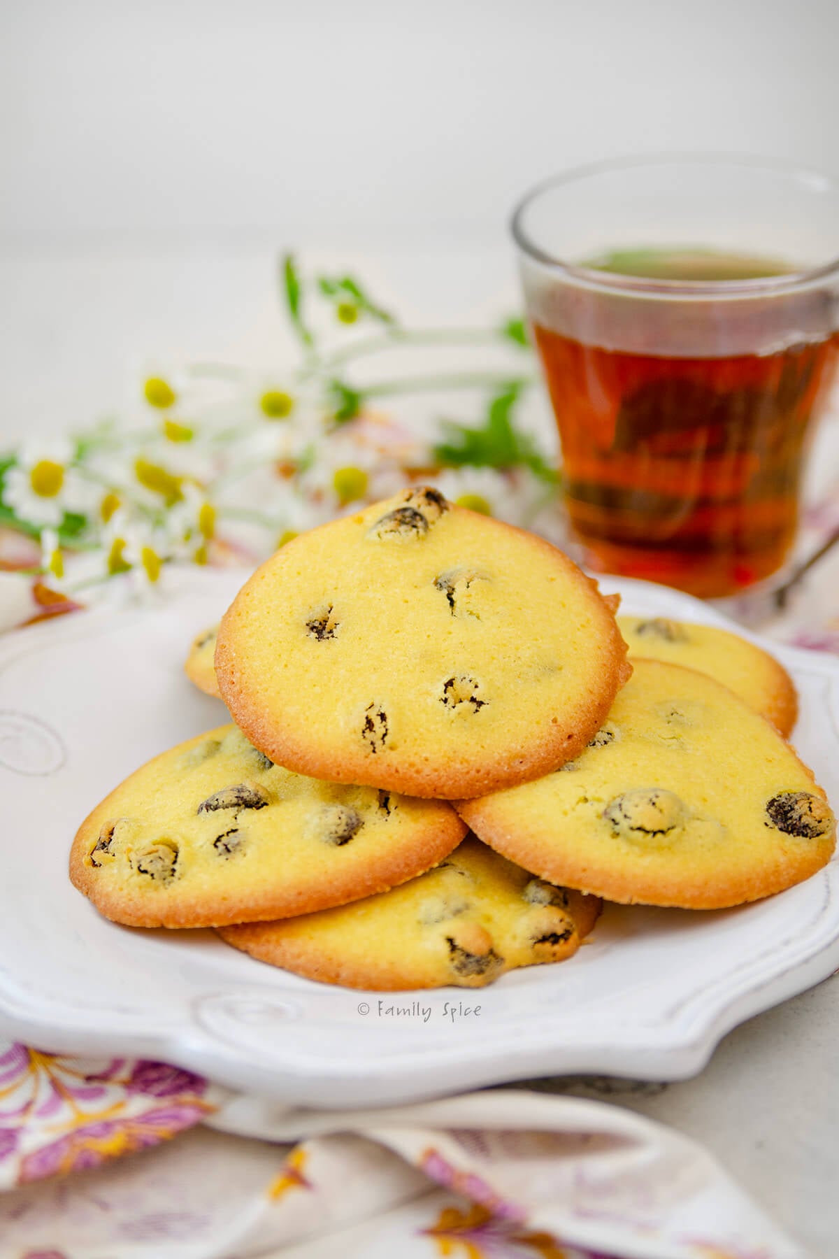 A white plate with Persian Saffron Raisin Cookies (shirini kishmishi) on it, plus flowers and cup of tea behind it.