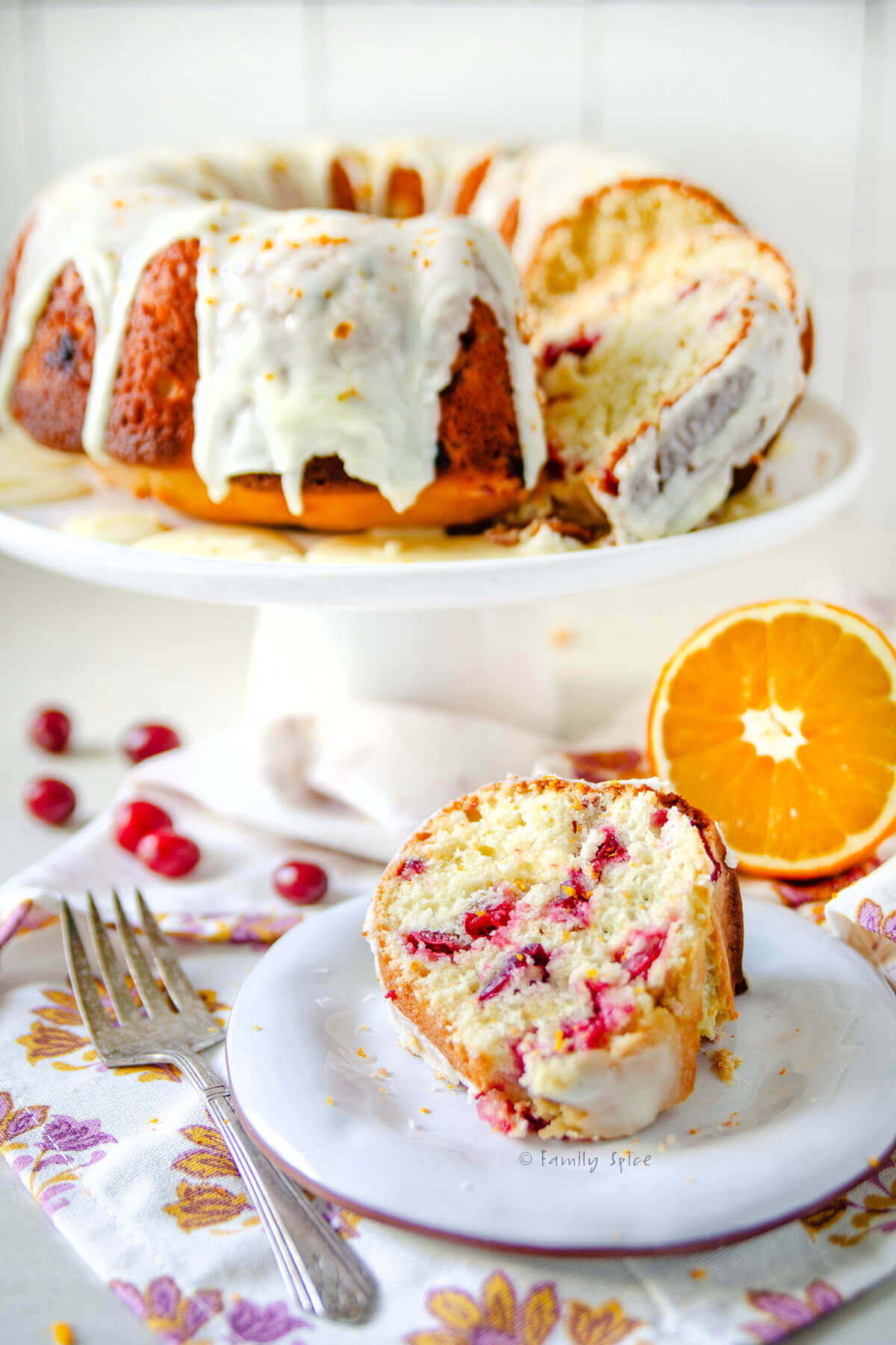 A slice of orange cranberry olive oil cake on a plate with the rest of the bundt cake behind it.