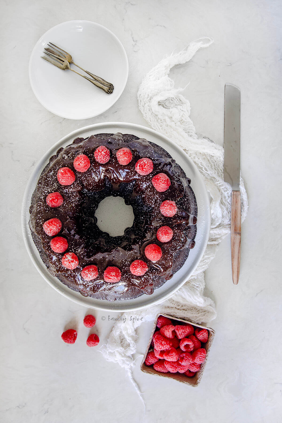 Top view of a chocolate bundt cake with a chocolate glaze and raspberries on a white platter with plates and serving knife next to it.