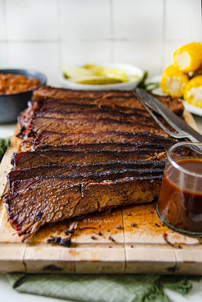 Side view of an oven baked brisket sliced up on a cutting board with corn, beans and pickle behind it.