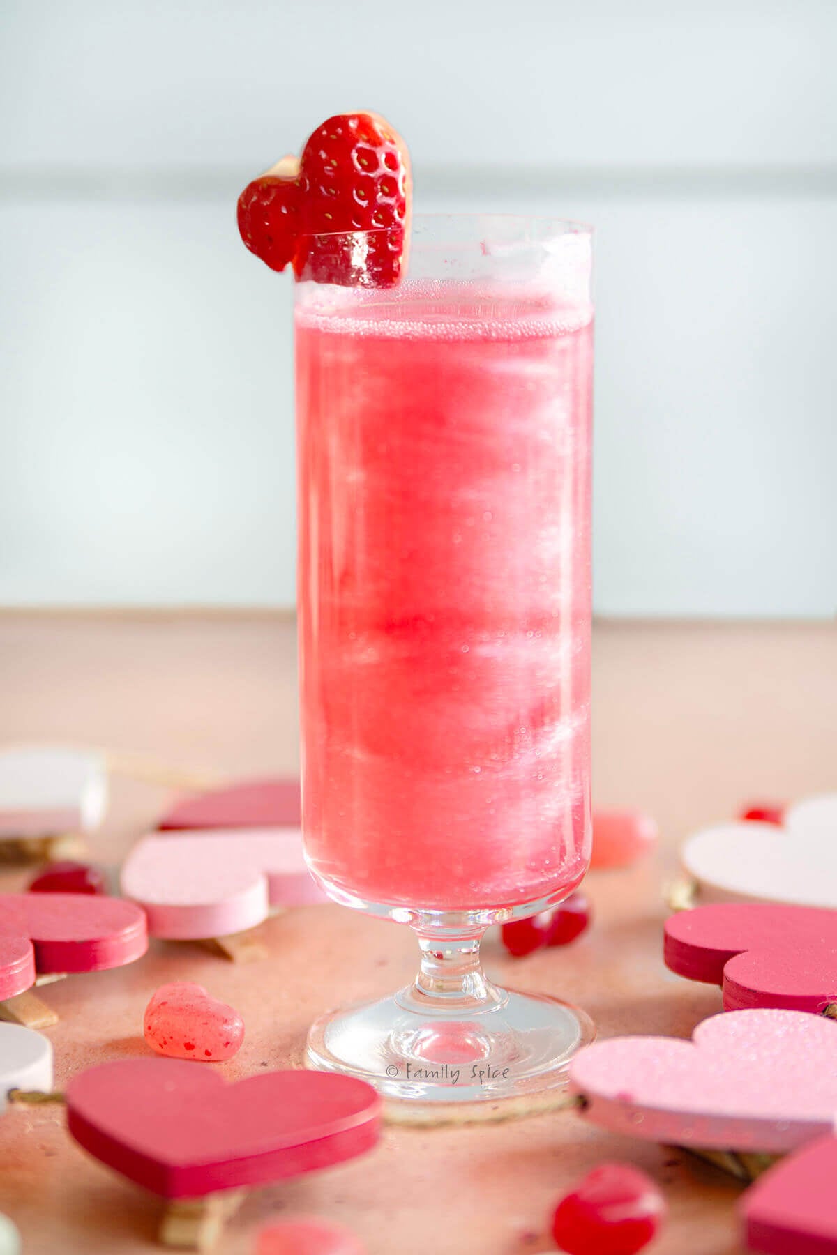Side view of a short stemmed champagne flute filled with shimmering pink mimosa garnished with a heart shaped strawberry.