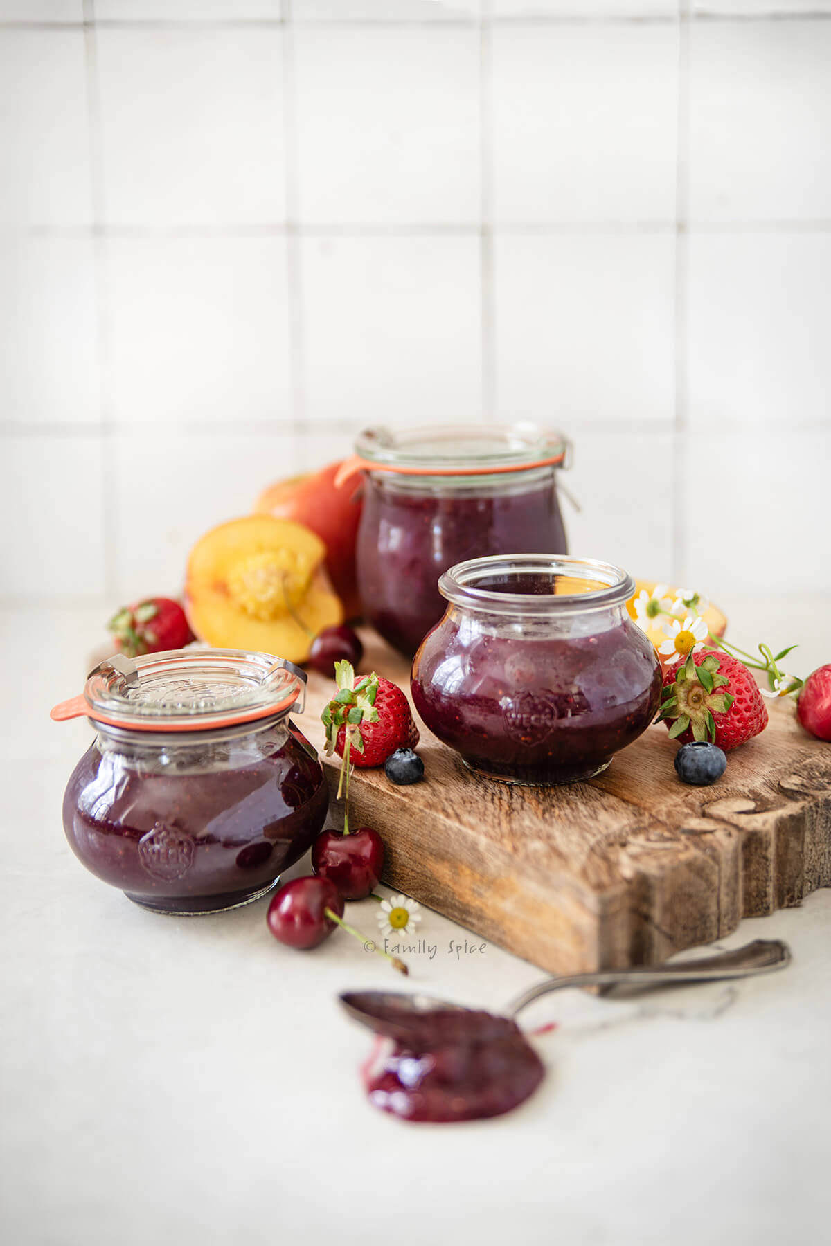 Three jars of mixed fruit jam on a cutting board with fruit around it.