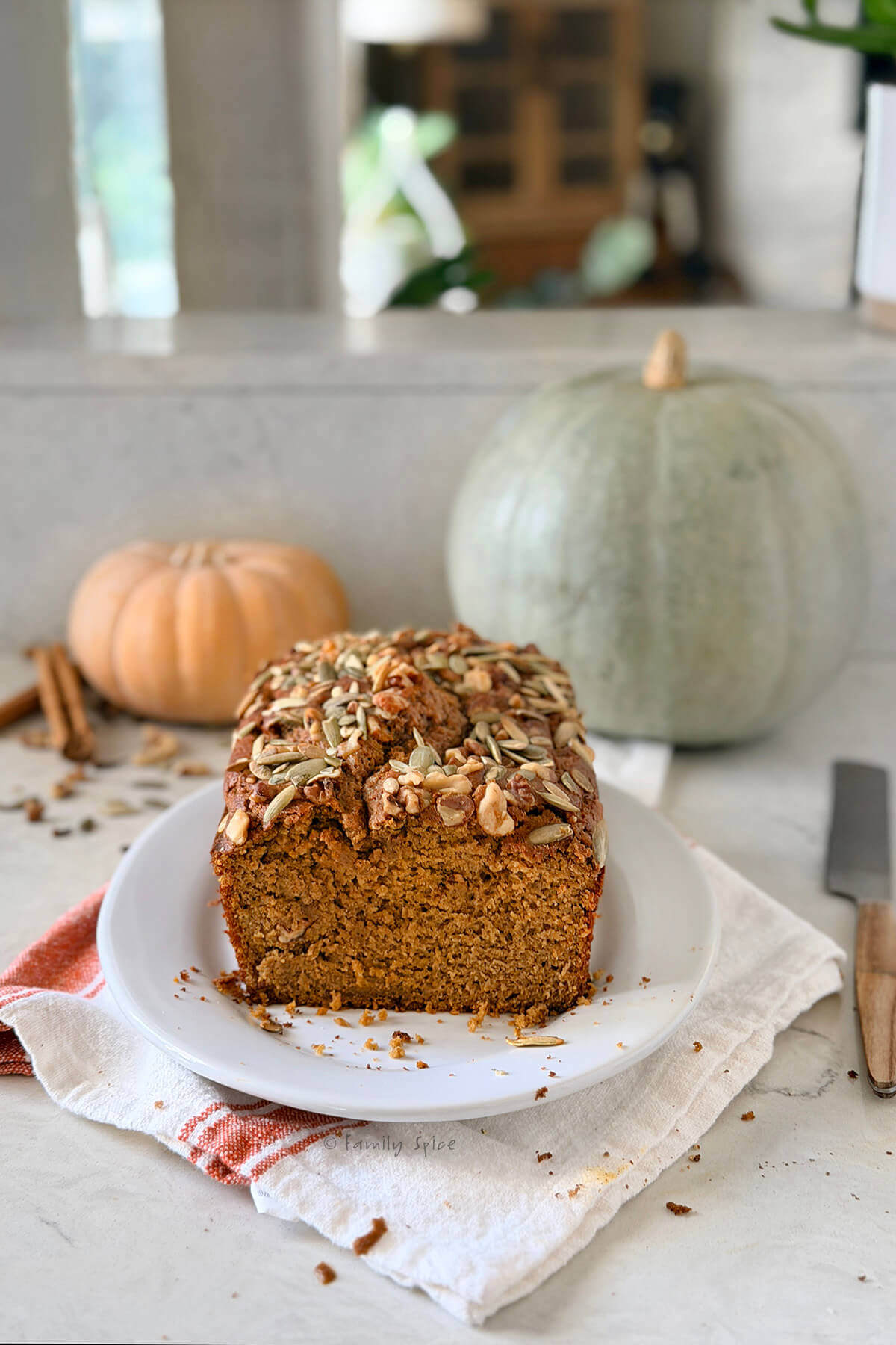 Side view of a loaf of olive oil pumpkin bread sliced open and two pumpkins behind it.