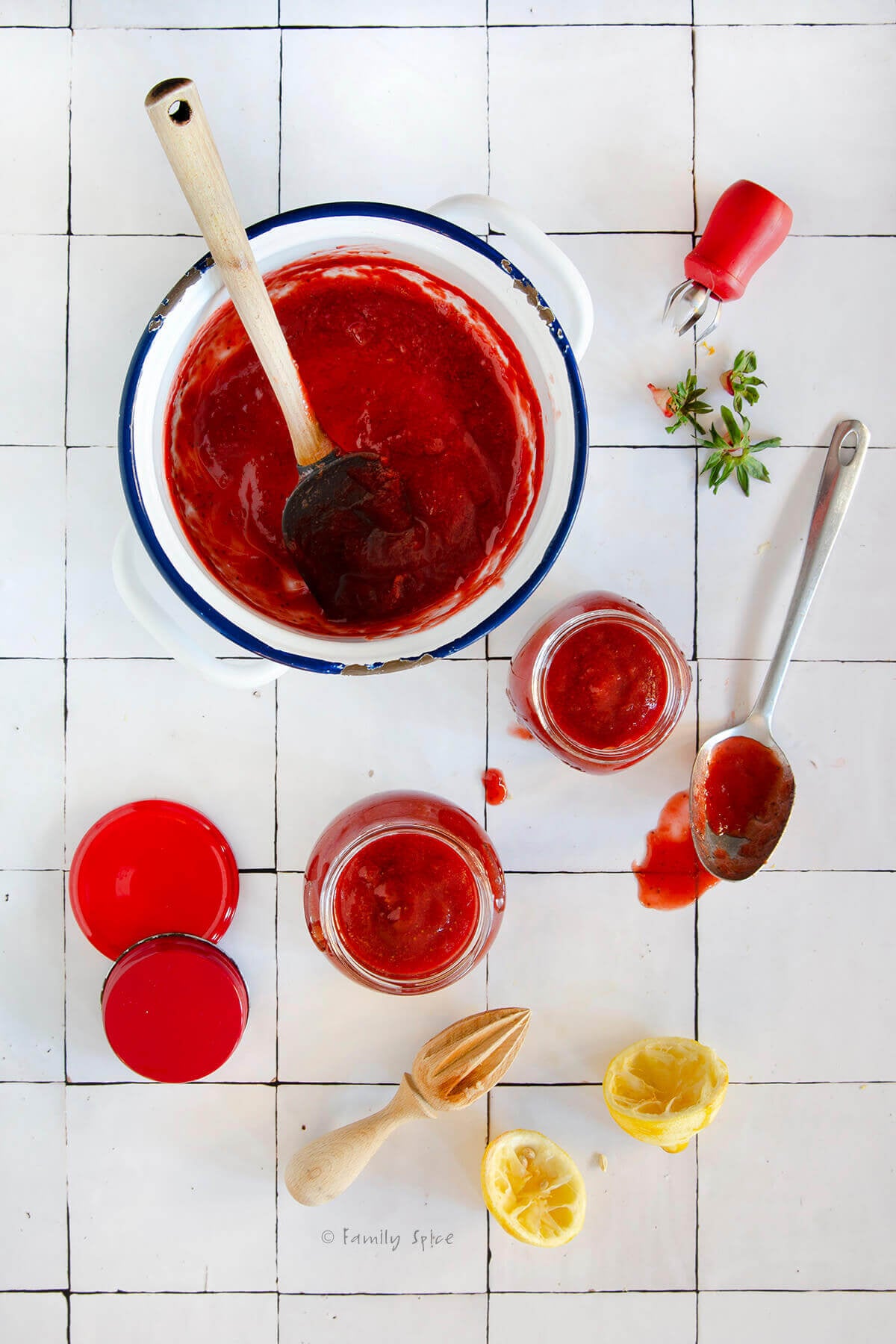 Top view of a pot with strawberry rhubarb jam and jars filled with jam alongside a spoon and lemons on a white surface.