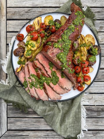 Top view of a round platter with grilled whiskey tri tip, topped with chimichurri and surrounded by grilled vegetables.