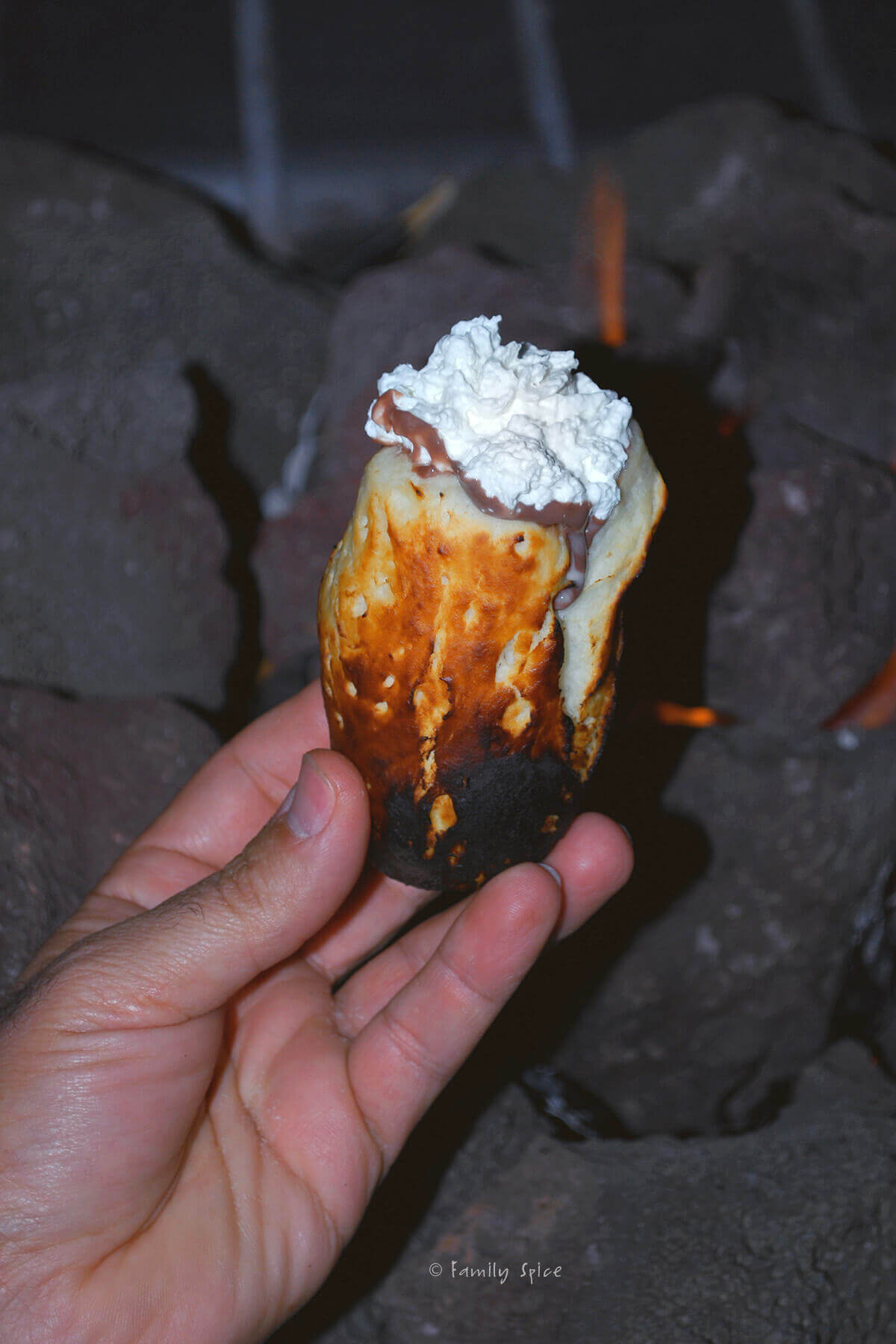 A hand holding a campfire eclair topped with whipped cream