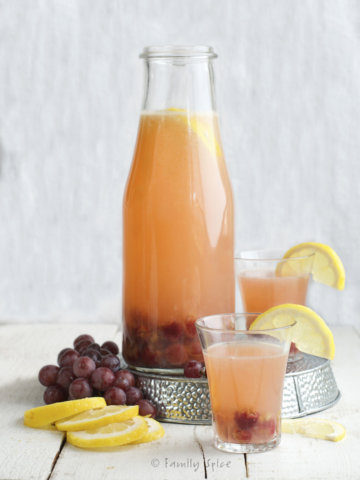 A large glass carafe of roasted grape lemonade with fresh grapes and lemons around it