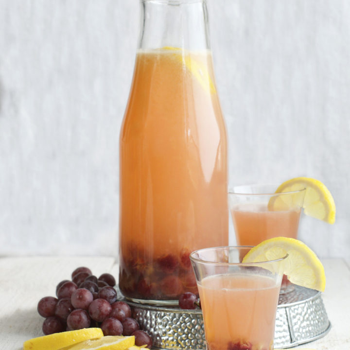 A large glass carafe of roasted grape lemonade with fresh grapes and lemons around it