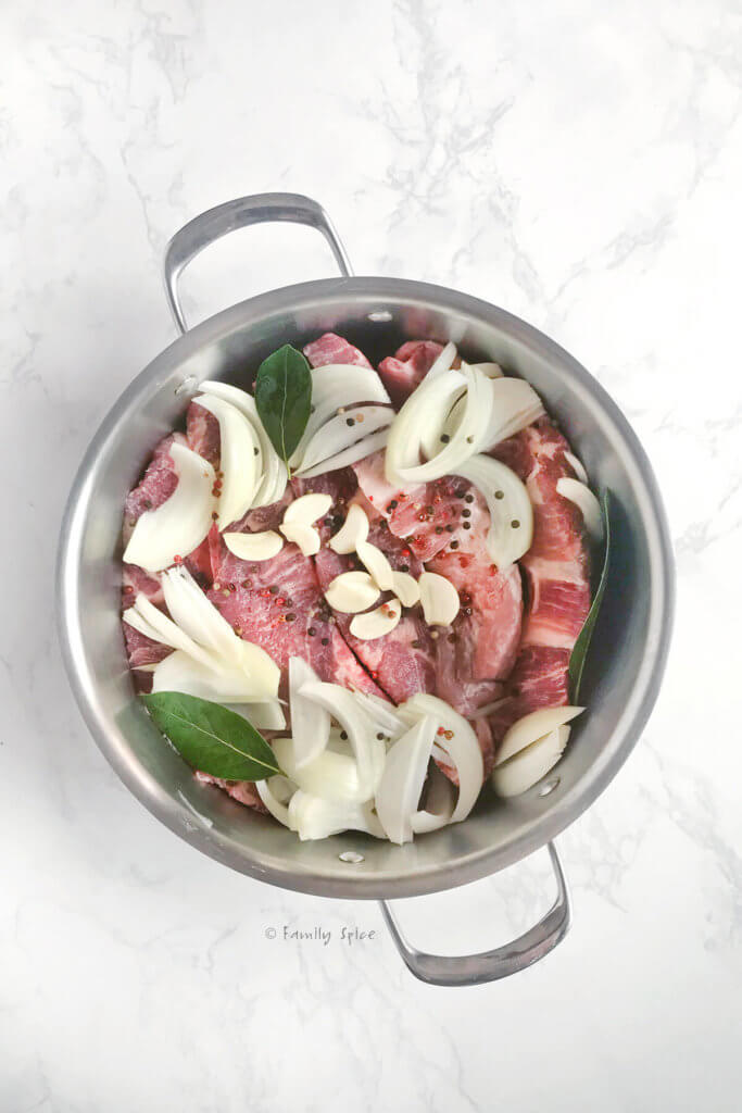 A large stainless steel pot with raw country style ribs, slices of onions and garlic and bay leaves in it