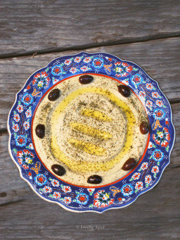 A blue ornate shallow bowl with hummus inside and garnished with olive oil and olives