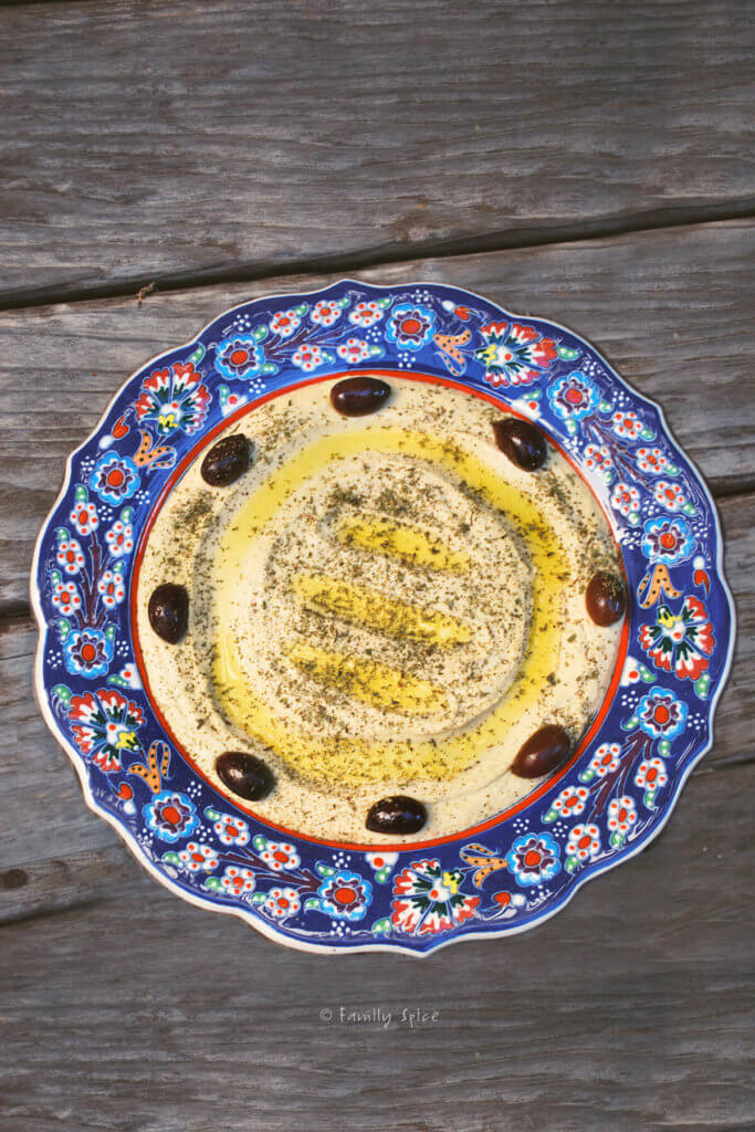 A blue ornate shallow bowl with hummus inside and garnished with olive oil and olives