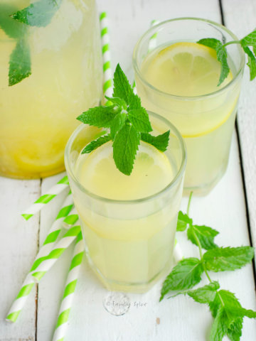 Closeup of two glasses of honey lemonade garnished with mint with straws and a pitcher of lemonade next to it