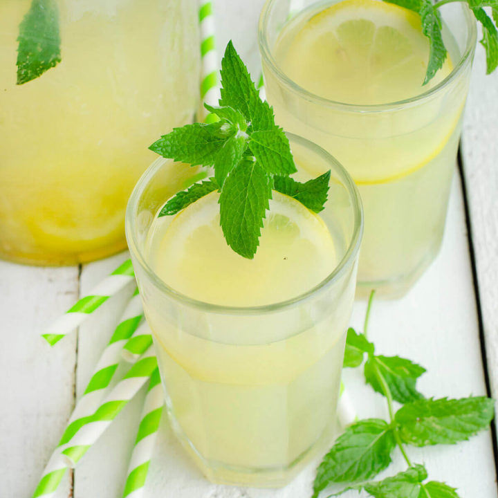 Closeup of two glasses of honey lemonade garnished with mint with straws and a pitcher of lemonade next to it