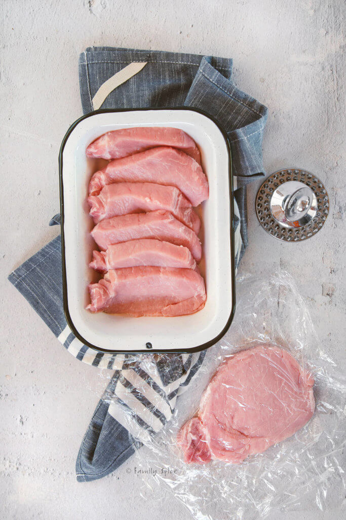 A pork loin cut into slices with one slice covered in plastic wrap and flattened into a cutlet