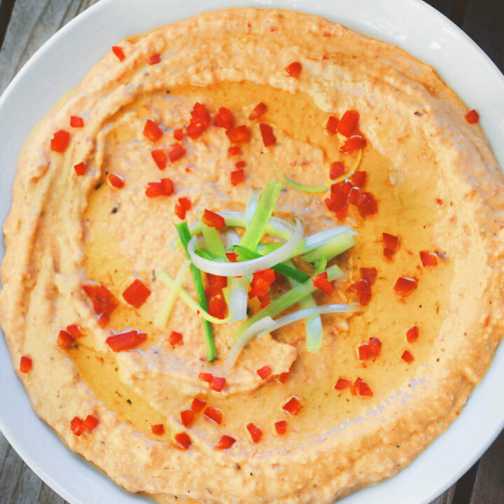 Overhead view of red pepper hummus garnished with green onions, chopped red pepper and olive oil