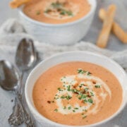 Closeup of a white bowl with shrimp bisque garnished with cream swirls, parsley and red pepper flakes with crispy bread sticks next to it