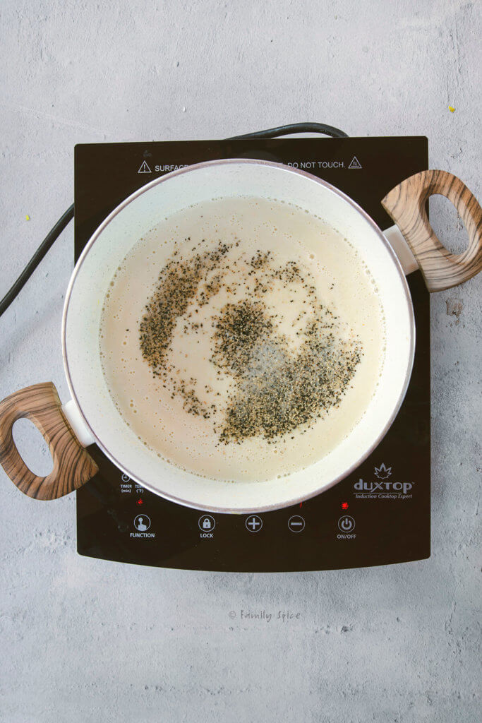 A pot on a cooktop with cream mixture simmering in it and seasoned with salt and pepper