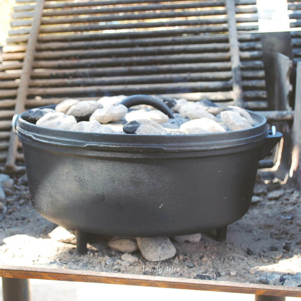 A cast iron Dutch oven in a campsite grill with coals under it and over it
