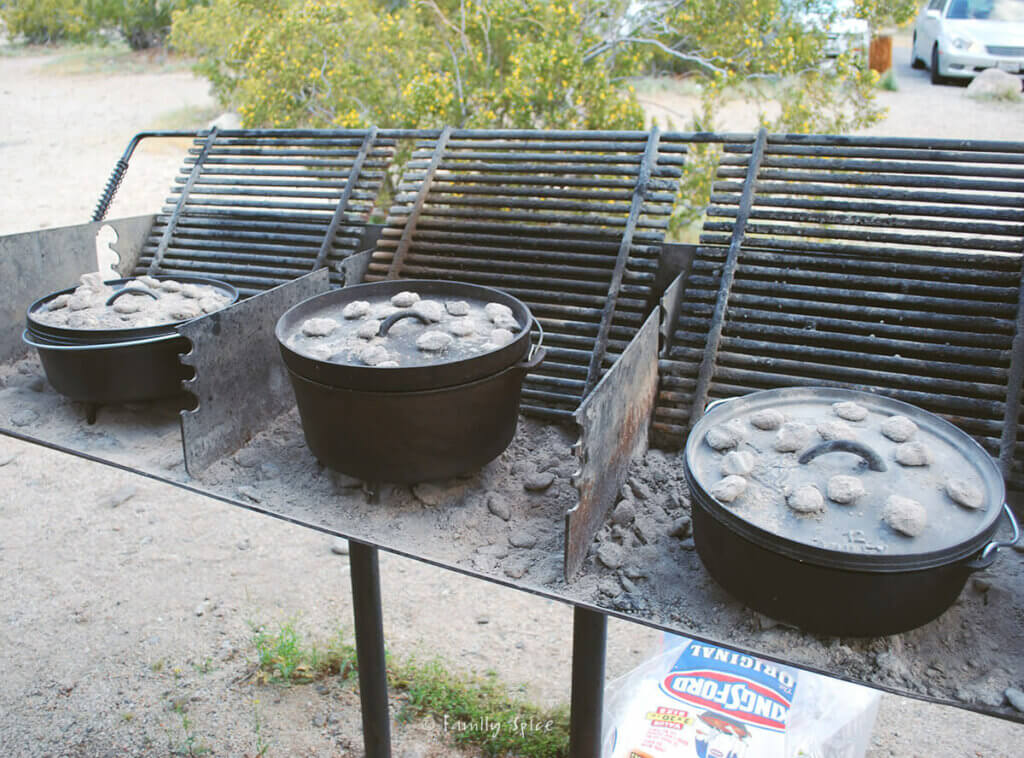 Three cast iron Dutch ovens in a campsite grill with coals under it and over it