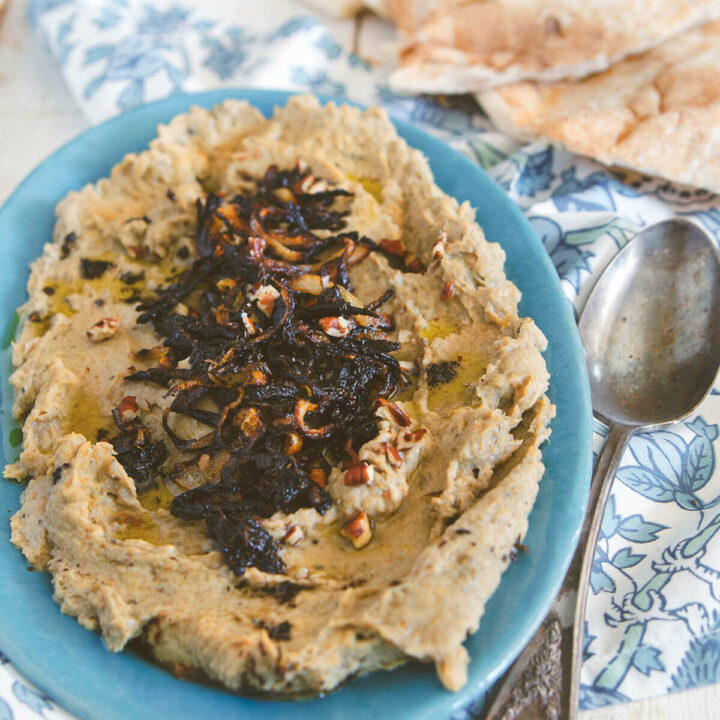 Closeup of an oval blue dish with kashkeh bademjan spread over it and garnished with caramelized onions, dried mint and olive oil