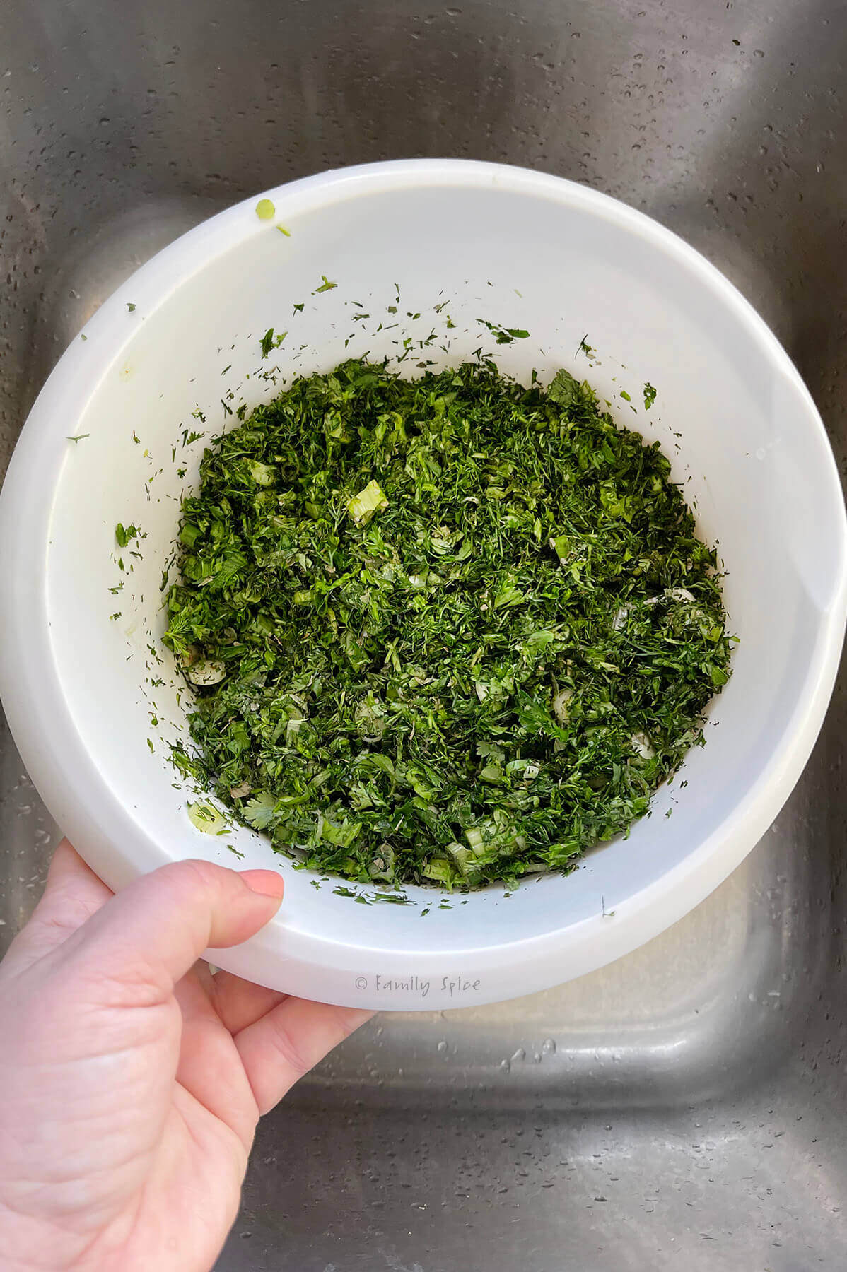 A hand holding a white bowl with chopped herbs in it