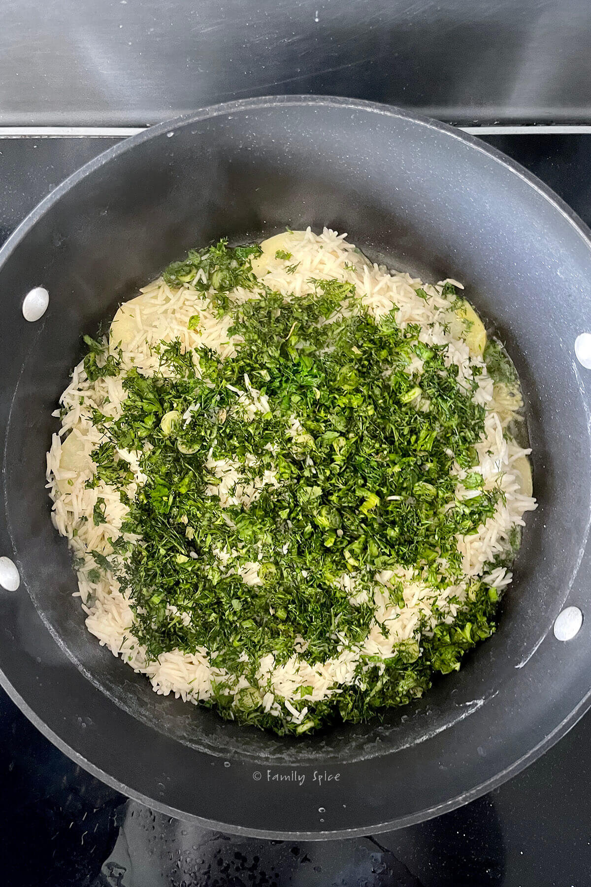 Top view of a non-stick pot with a layer of chopped herbs over a layer of parboiled rice