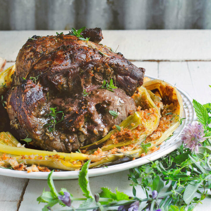 Butterflied leg of lamb stuffed with mushrooms and herbs on a platter with grilled leeks and fresh herbs