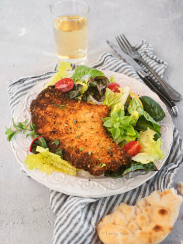 Closeup shot of one pork Milanese cutlet on a plate with greens, lemon wedge and tomatoes with a glass of white wine