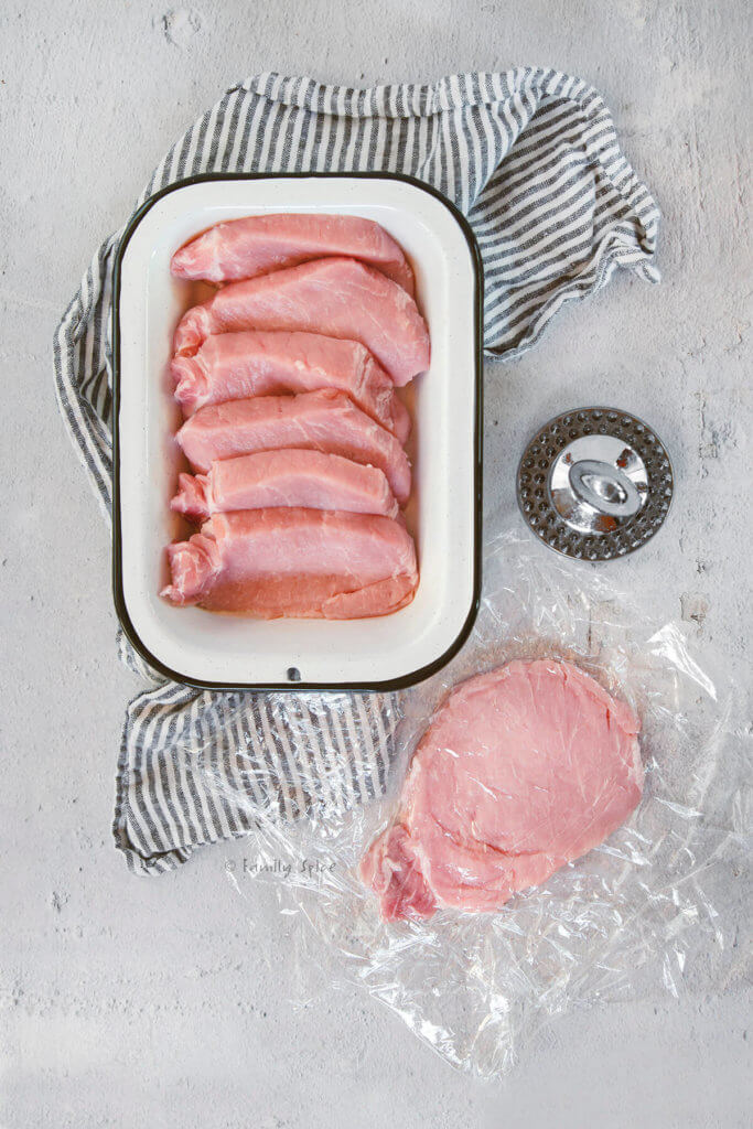 A pork loin cut into slices with one slice covered in plastic wrap and flattened into a cutlet