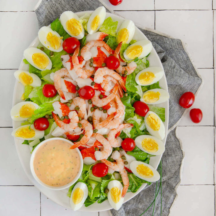 Top view of an oval platter with shrimp and crab louie salad arranged neatly and a bowl of louie dressing next to it