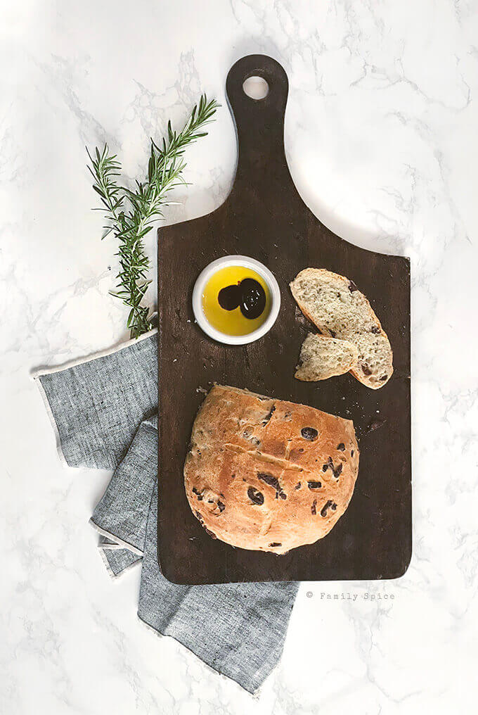 A ball of rosemary olive bread on a dark wood cutting board sliced with a small bowl of olive oil and vinegar by FamilySpice.com