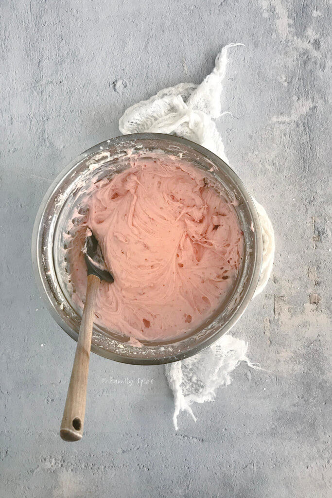 A mixing bowl filled with homemade strawberry frosting with a rubber scraper
