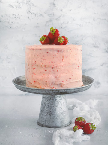 Side view of a 3 layered strawberry cake with strawberry frosting and topped with fresh strawberries on a rustic metal cake stand