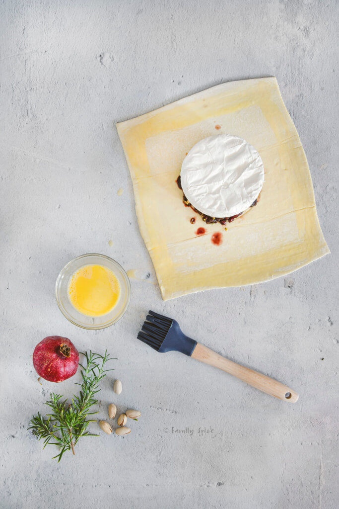 A wheel of brie on top of fruits and nuts on a sheet of raw puff pastry on a concrete work surface with egg wash and brush next to it