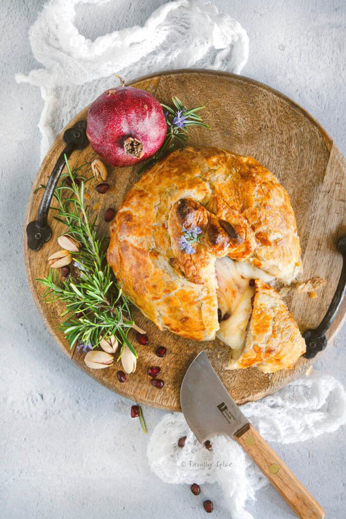 Top view of a baked brie wrapped in puff pastry and stuffed with pomegranate and nuts cut open on a round wooden tray with rosemary and pomegranate next to it