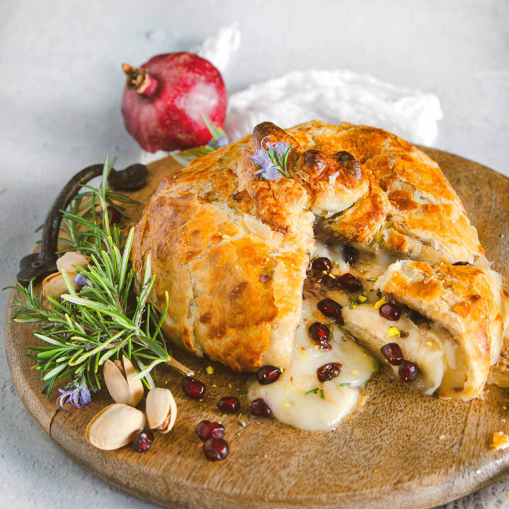 Baked brie wrapped in puff pastry and stuffed with pomegranate and nuts cut open on a round wooden tray