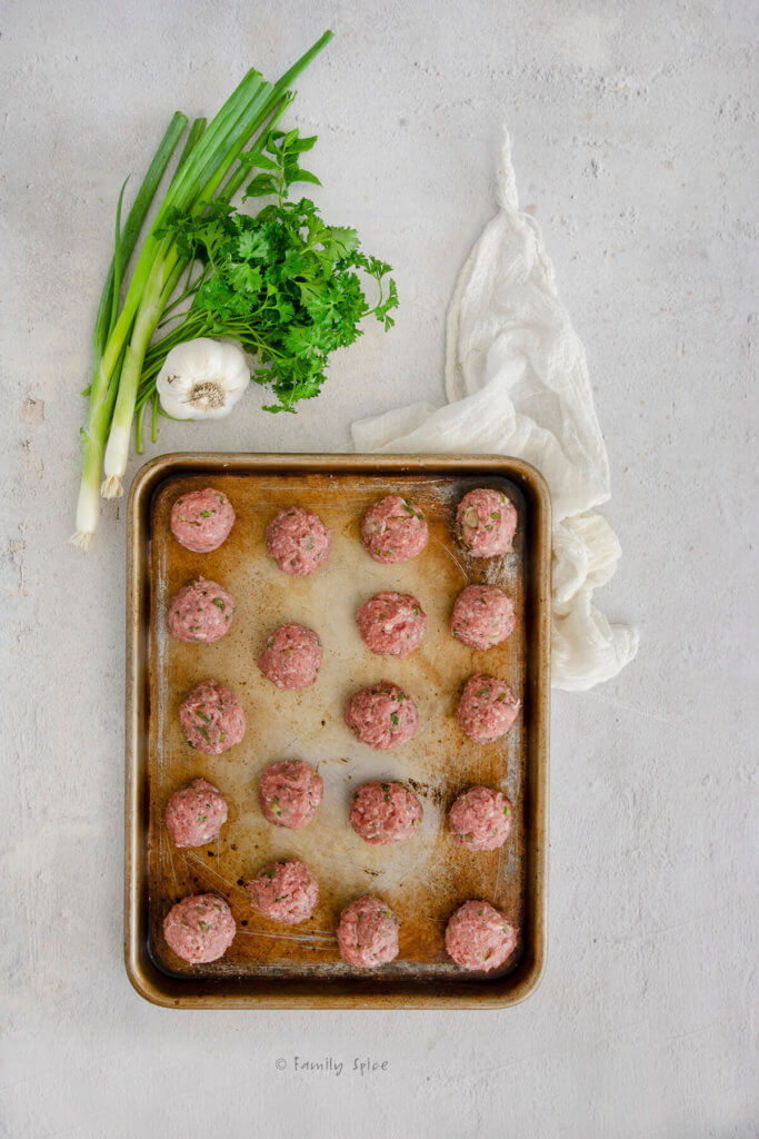 Raw lamb meatballs on a small baking sheet with fresh herbs and garlic next to it