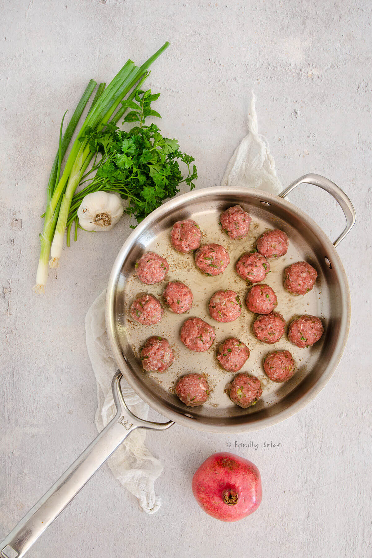 Browning lamb meatballs in a stainless frying pan with fresh herbs and garlic next to it
