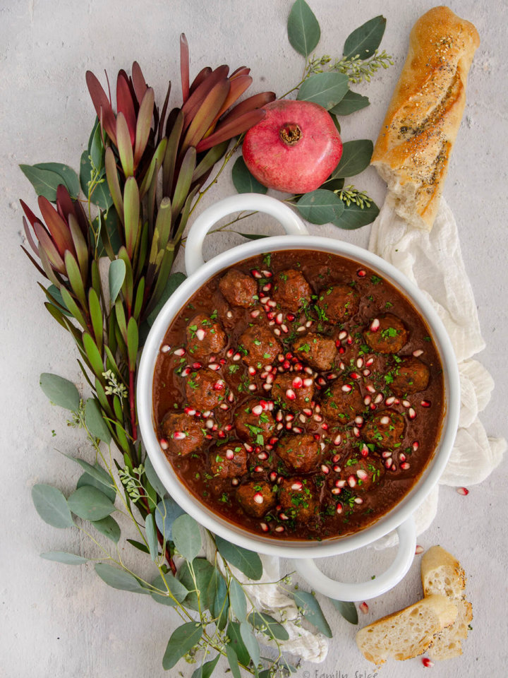 A white serving bowl with lamb meatballs in tomato pomegranate sauce with greenery and baguette around it