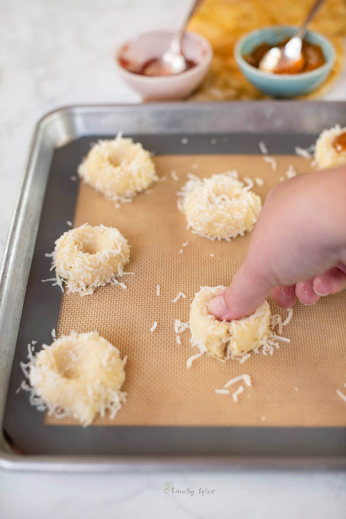 A hand pressing a thumb into cookies rolled in coconut on a baking sheet