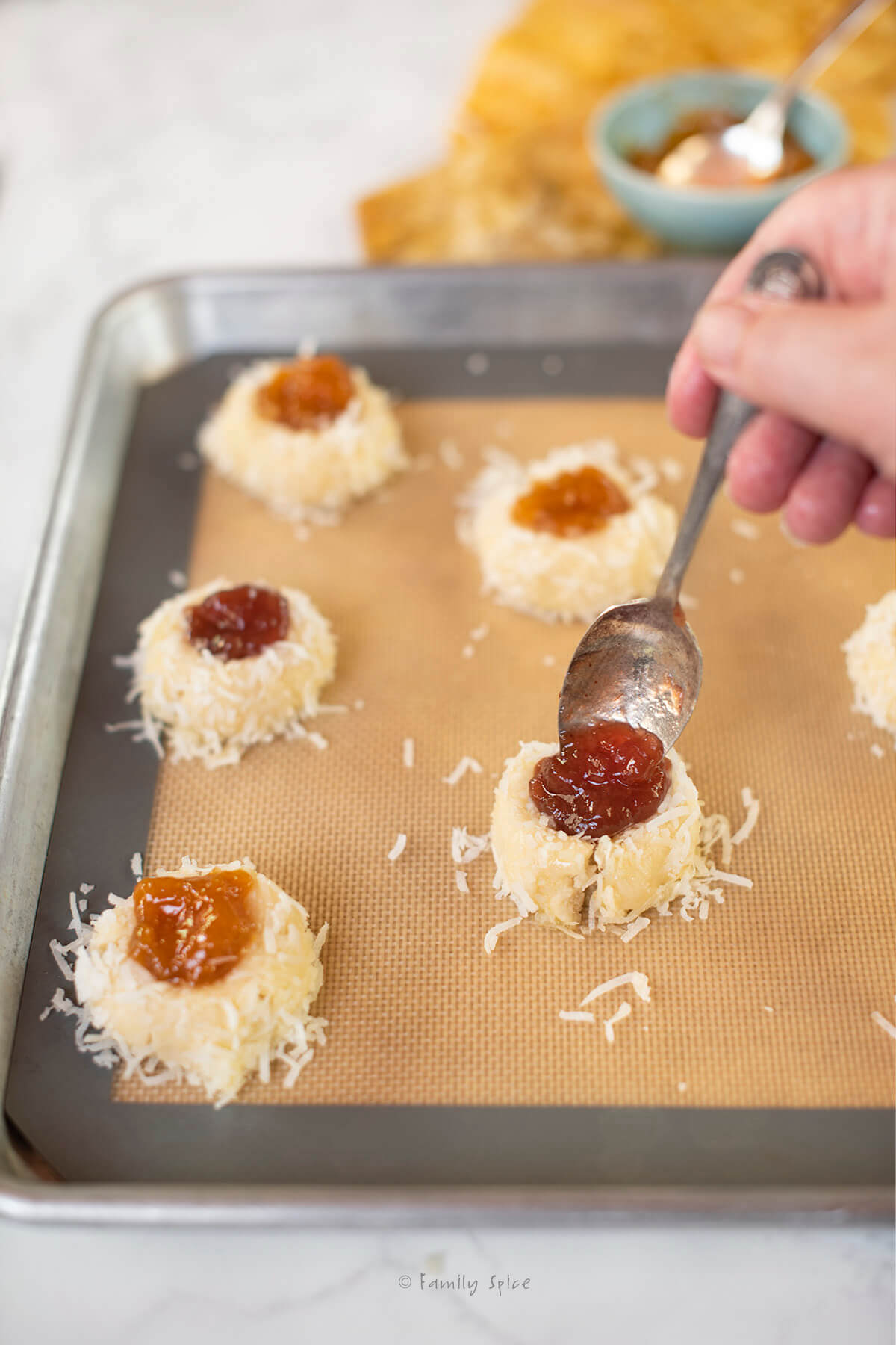 A hand spooning jam into well of coconut thumbprint cookies on a baking sheet