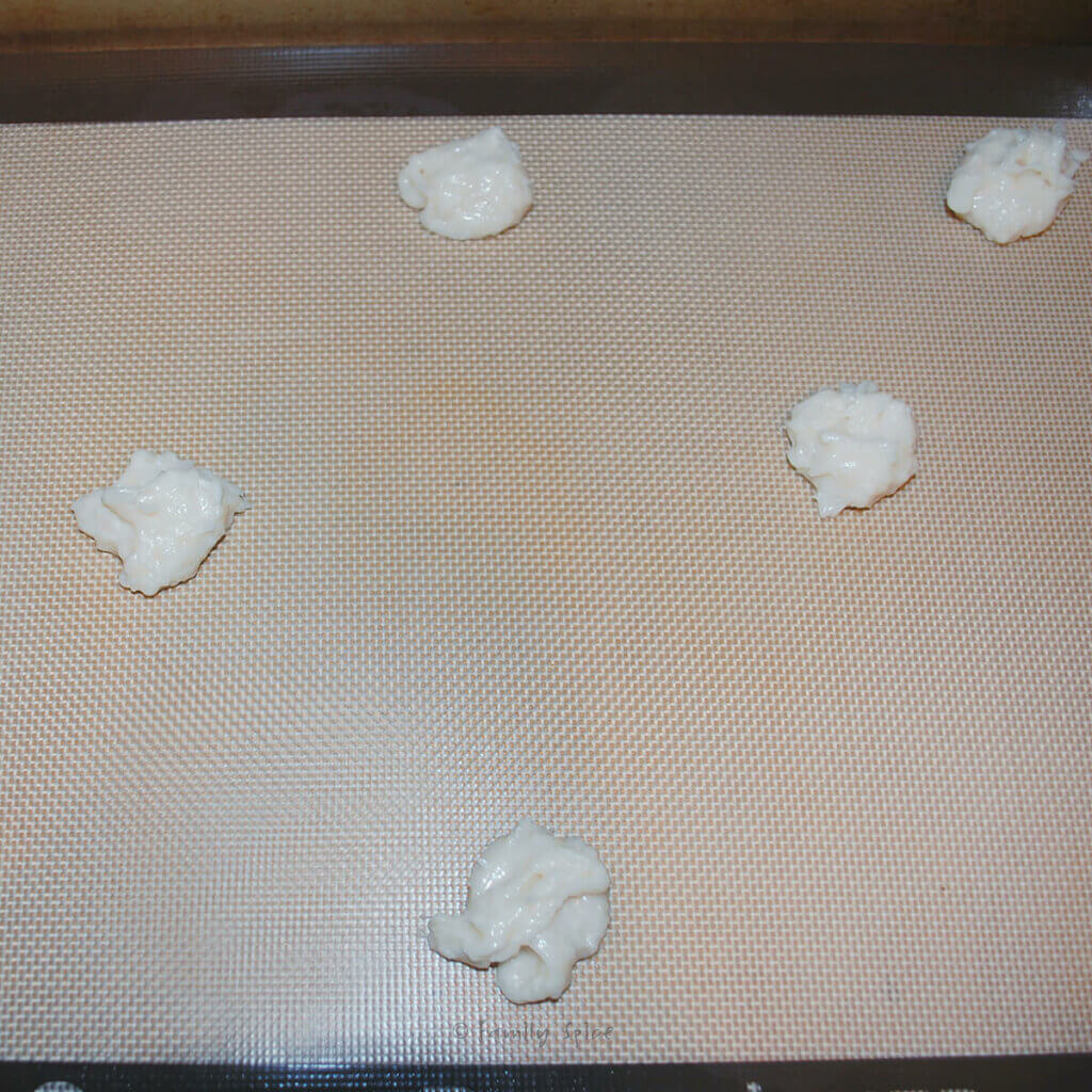 A baking sheet lined with silicone baking mat with teaspoons of coconut thin cookie batter on it