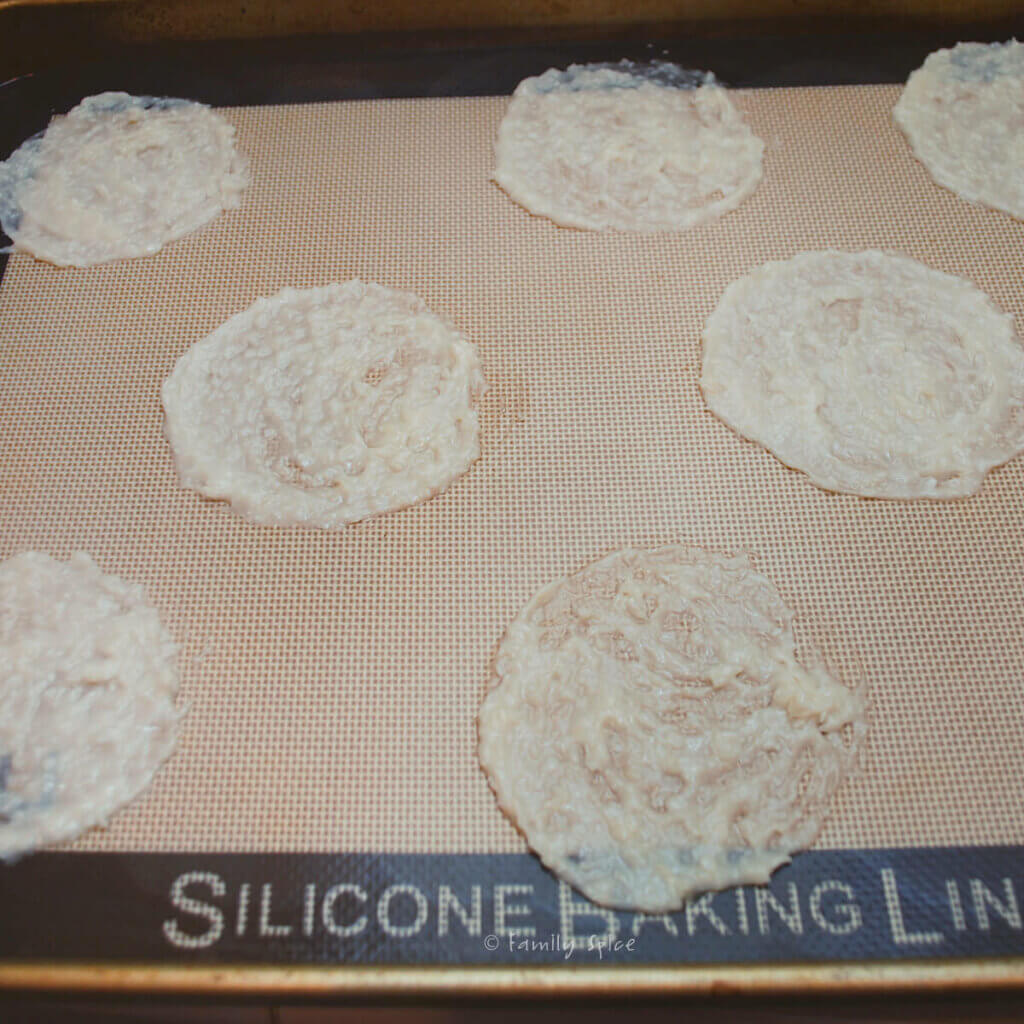 A baking sheet lined with silicone baking mat with coconut thin cookie batter spread thin on it