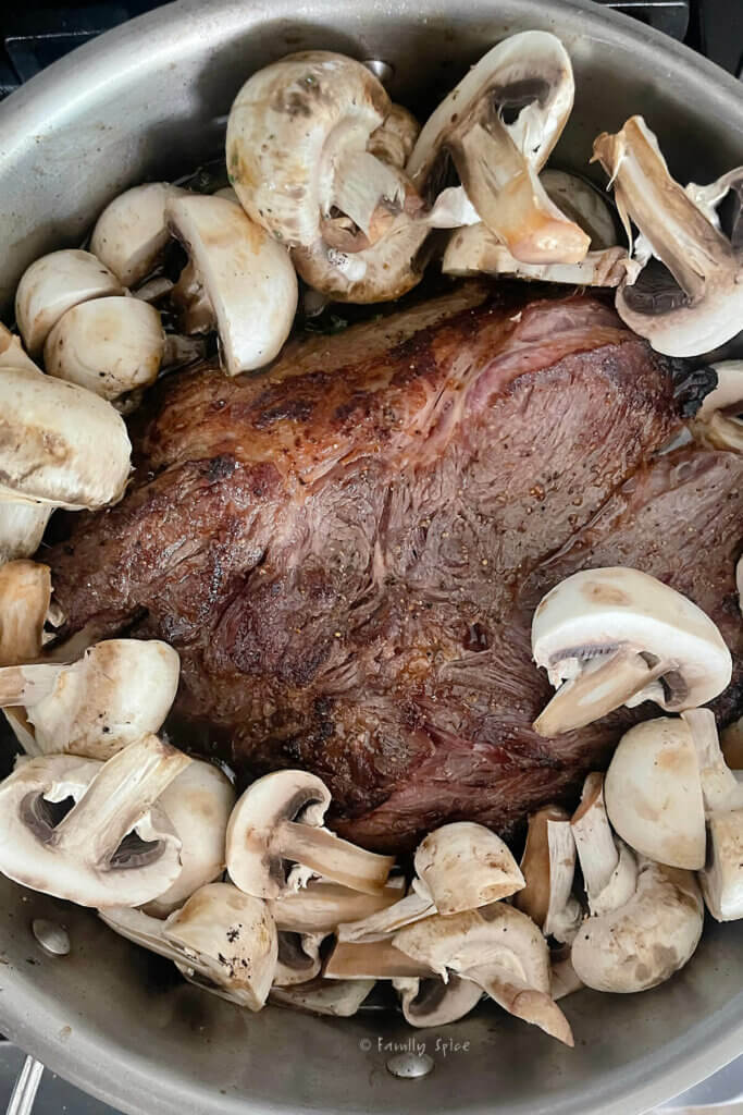 Top view of a stainless pot with a braised chuck roast in it and slices of raw mushrooms around it