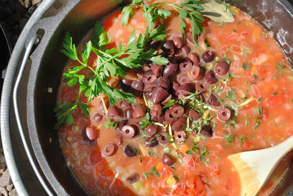 Adding herbs and chopped olives to tomato sauce simmering in a dutch oven outdoors